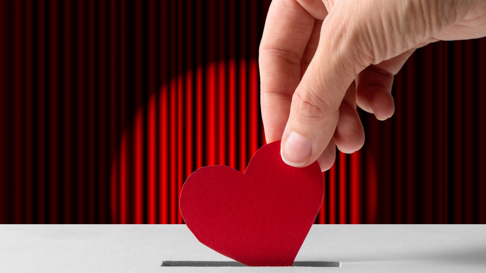 A photograph of a stage curtain. A large hand is coming above teh stage and posting a red paper heart into a donations box.